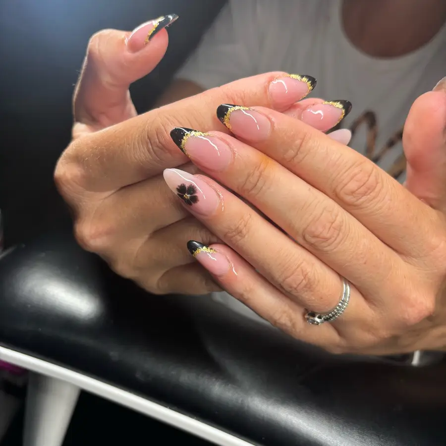 Almond acrylics with a nude base, black French tips edged in gold foil and one nail with a small black flower.