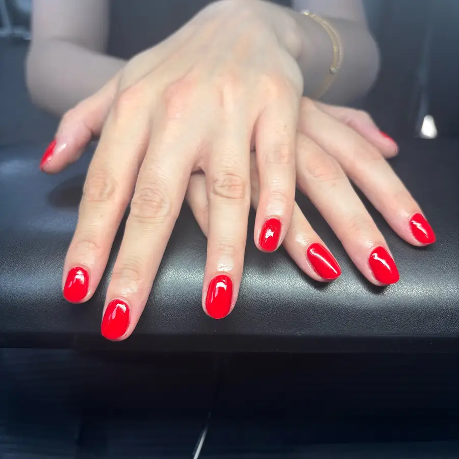 Short rounded nails in glossy bright red gel, hands resting on a black chair arm.
